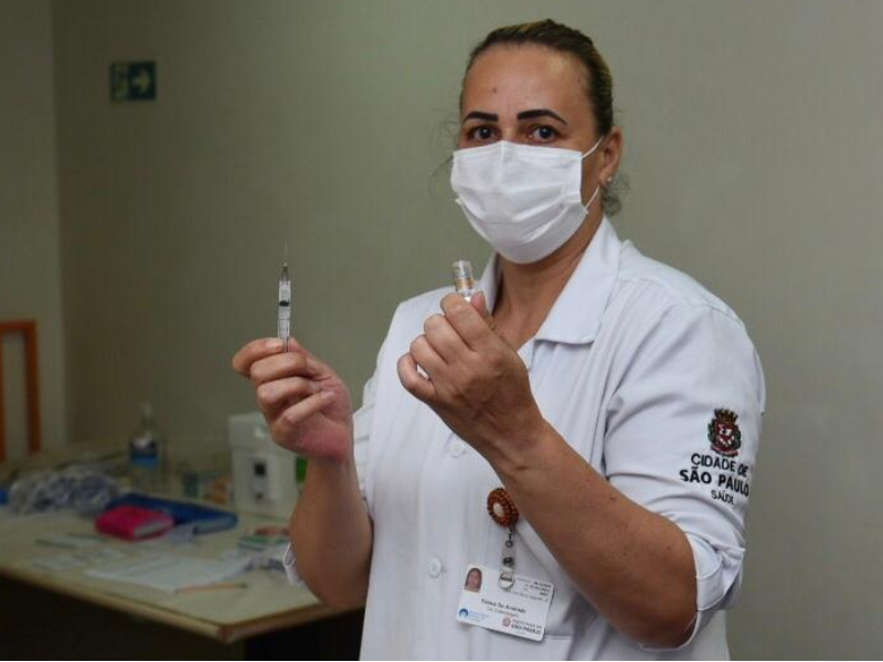 Uma profissional da saúde segura uma seringa numa das mãos e o frasco da vacina na outra mão. Atrás dela, à esquerda, há uma mesa com alguns objetos 